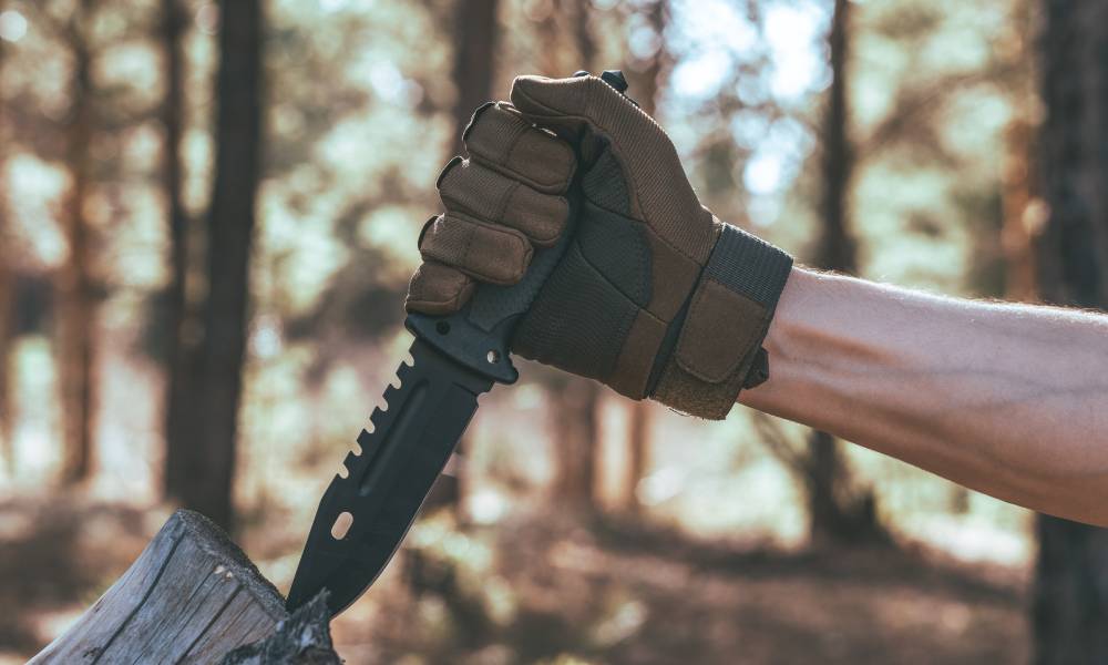A hand holding a knife while sticking it into the end of a log. The hand has a glove on and there's a forrest in the background.