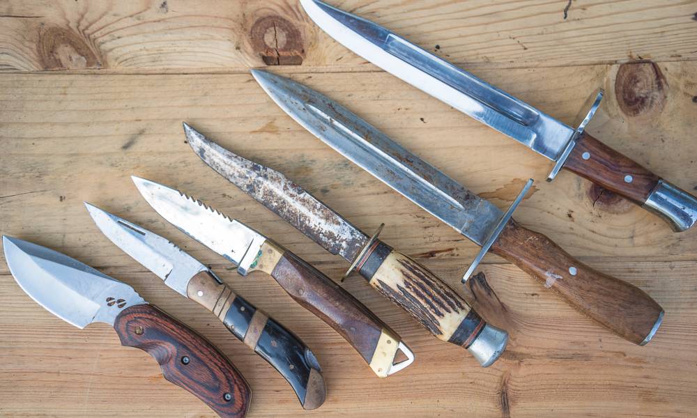 A collection of six hunting knives lying next to each other on a wooden table. They're in order of smallest to largest.