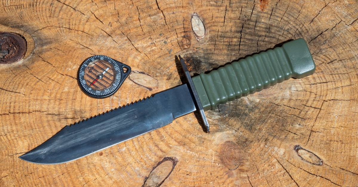 A survival knife with a green handle lying on top of a wooden stump. There is a small compass sitting next to it.
