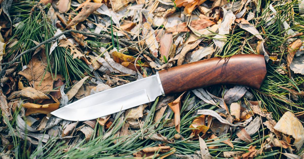 A hunting knife lying on a bed of leaves and twigs. There are also some pine needles in the foreground.