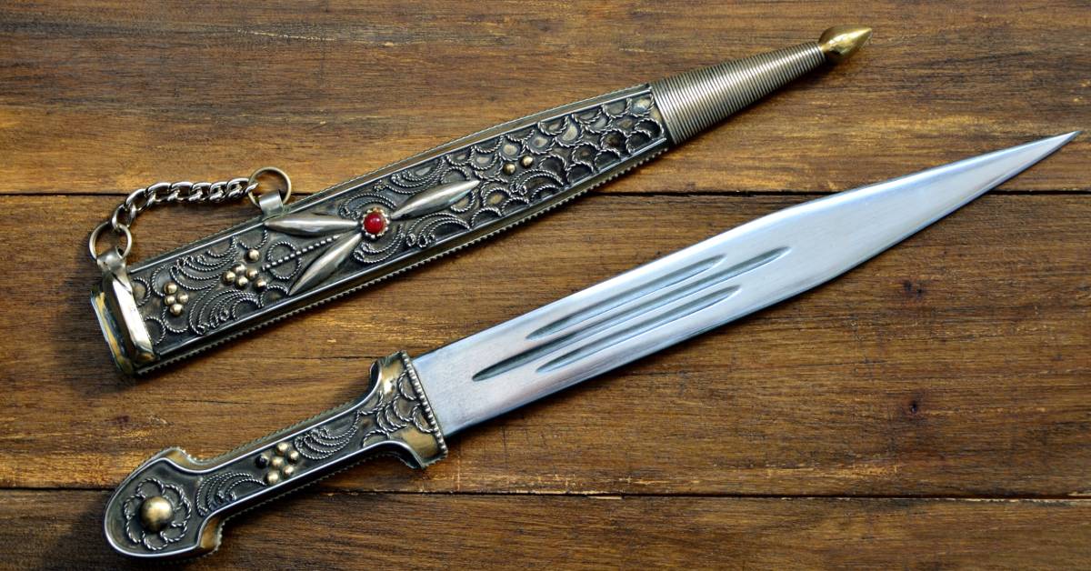 A old dagger lying on a wood table. The dagger's sheath is next to it and both are intricately detailed.