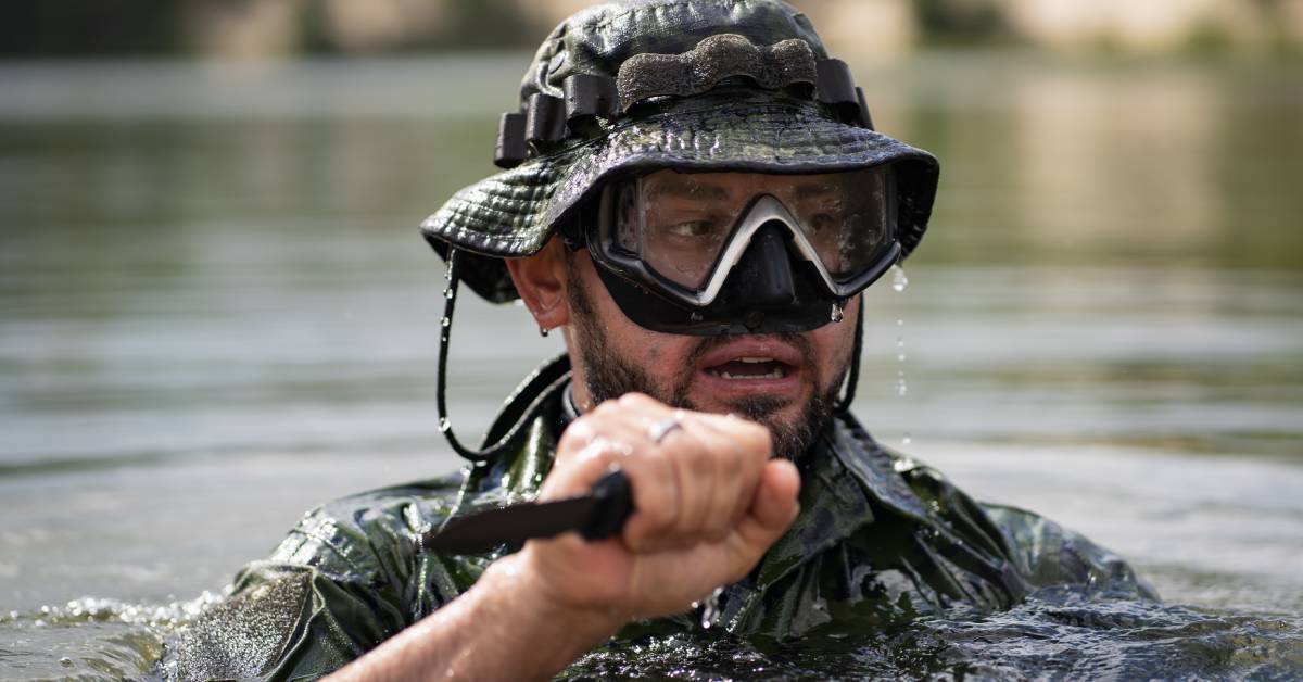 A guy wearing underwater combat gear coming out of the water. He is holding a combat knife in his right hand.